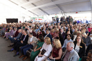 A large audience sits attentively under a white tent, facing a stage where media personnel are positioned.