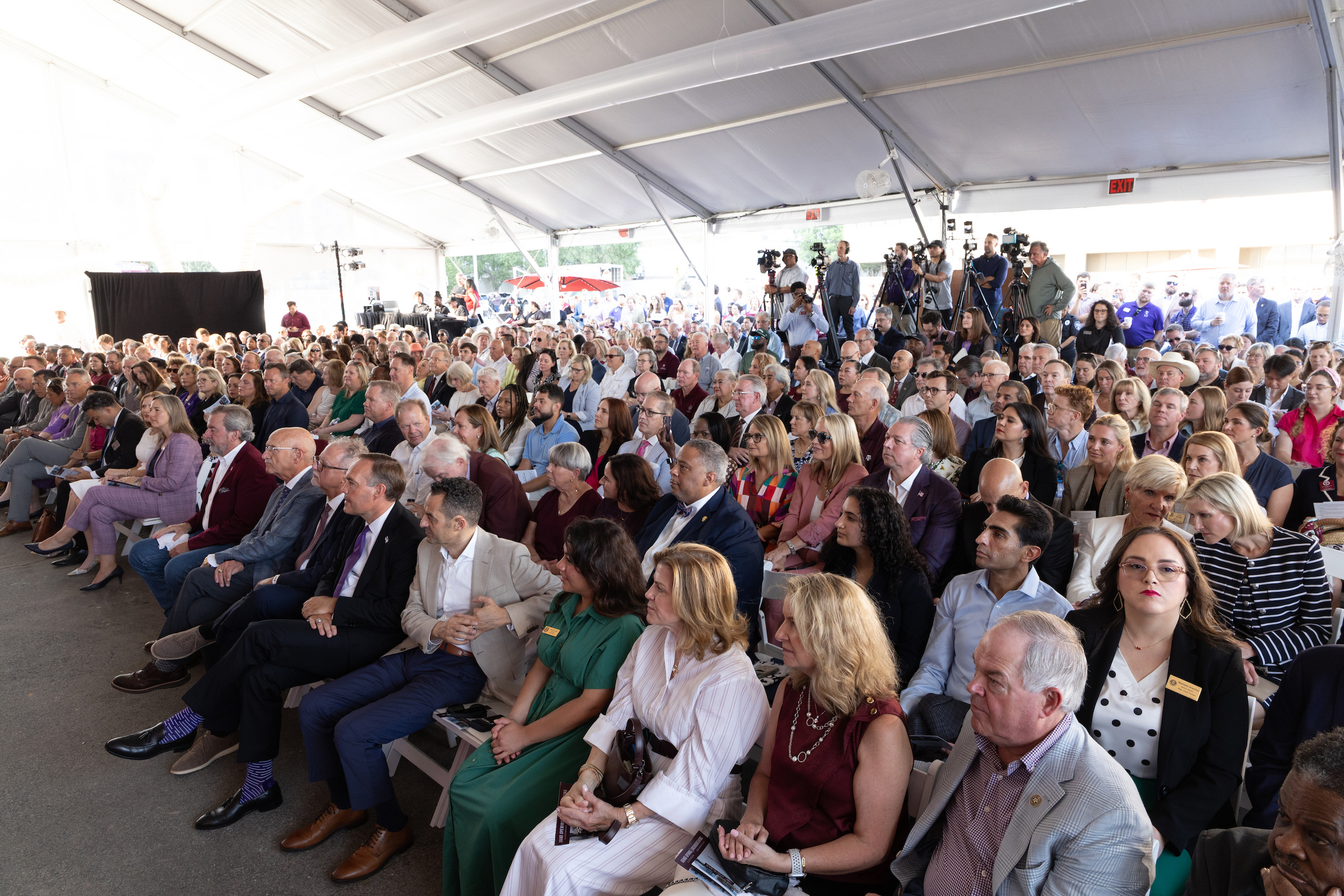 A large audience sits attentively under a white tent, facing a stage where media personnel are positioned.