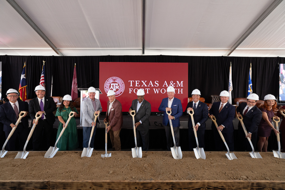 A group of individuals in hard hats stand together at a groundbreaking ceremony for Texas A&M Fort Worth, each holding a ceremonial shovel over freshly turned soil.