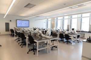 A modern laboratory with rows of workstations, microscopes, and a large screen displaying cellular imagery.