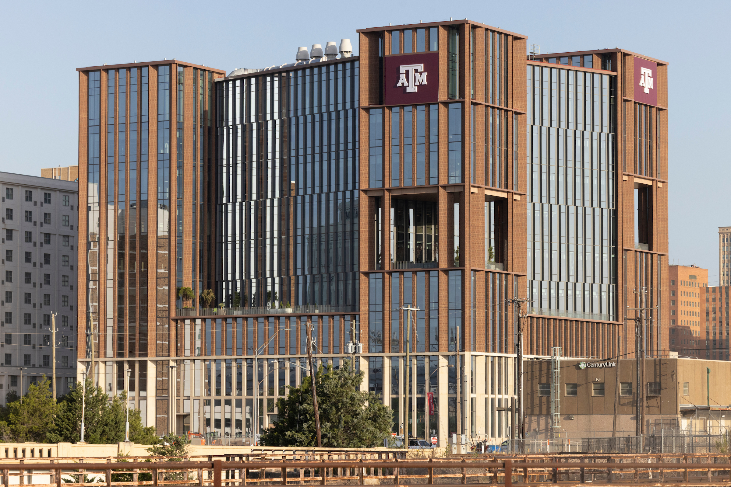A modern multi-story building with prominent Texas A&M University logos stands against a clear sky, surrounded by urban structures and greenery.